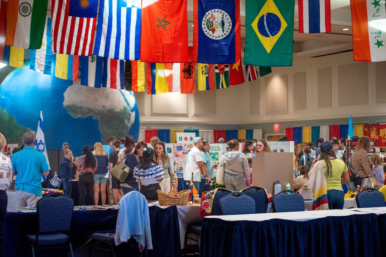 Busy international fair with a large globe, flags from around the world hanging from the ceiling, and people milling around tables.